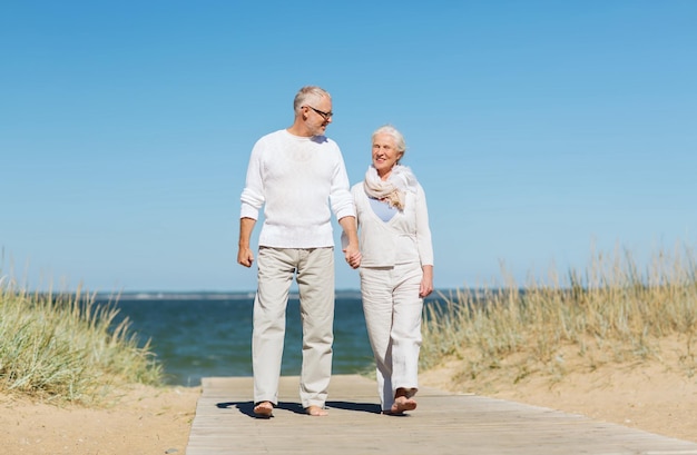 família, idade, viagens, turismo e conceito de pessoas - feliz casal sênior de mãos dadas e caminhando na praia de verão