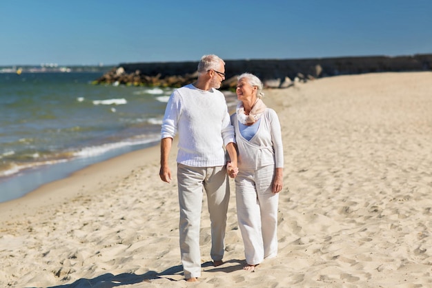 família, idade, viagens, turismo e conceito de pessoas - feliz casal sênior de mãos dadas e caminhando na praia de verão