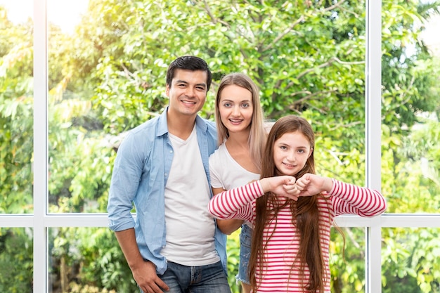 Familia, incluido el padre, la madre y la hija, de pie y abrazándose frente a la ventana de vidrio en casa mirando la cámara