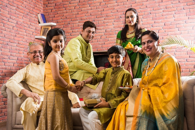 Familia india celebrando el festival Raksha Bandhan o Rakhi en casa mientras usa ropa tradicional y se sienta en el sofá