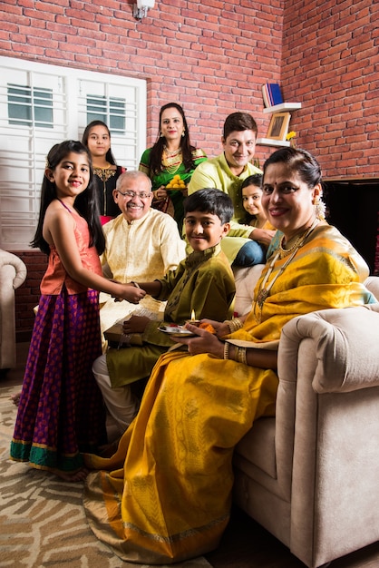 Familia india celebrando el festival Raksha Bandhan o Rakhi en casa mientras usa ropa tradicional y se sienta en el sofá