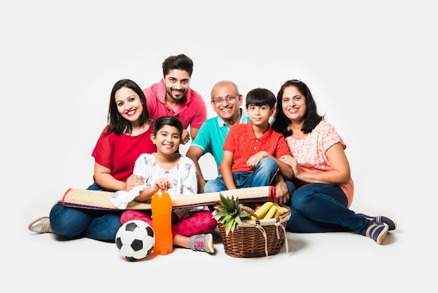 Familia india disfrutando de un picnic en el interior - Varias generaciones de familias asiáticas sentadas sobre fondo blanco con canasta de frutas, tapete, fútbol y bebidas. enfoque selectivo