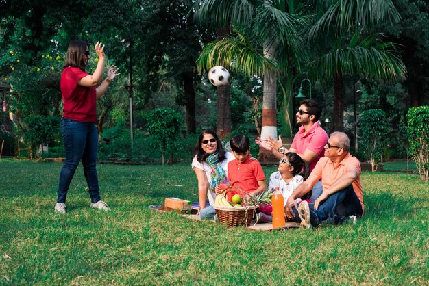 Familia india disfrutando de un picnic: varias generaciones de familias asiáticas sentadas sobre el césped o la hierba verde en el parque con canasta de frutas, tapete y bebidas. enfoque selectivo