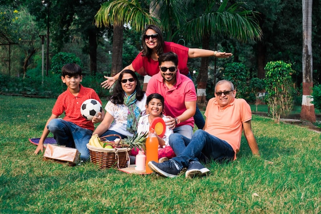 Familia india disfrutando de un picnic: varias generaciones de familias asiáticas sentadas sobre el césped o la hierba verde en el parque con canasta de frutas, tapete y bebidas. enfoque selectivo