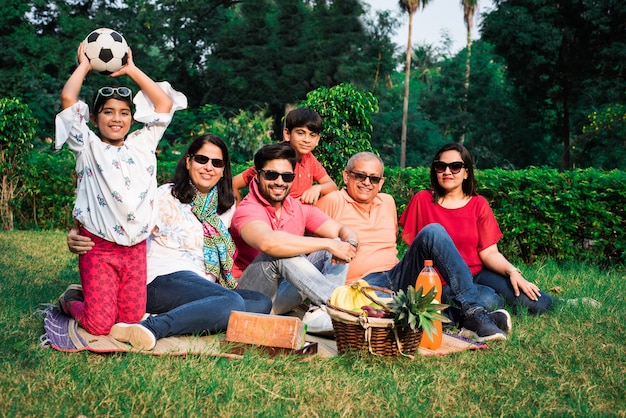 Familia india disfrutando de un picnic: varias generaciones de familias asiáticas sentadas sobre el césped o la hierba verde en el parque con canasta de frutas, tapete y bebidas. enfoque selectivo