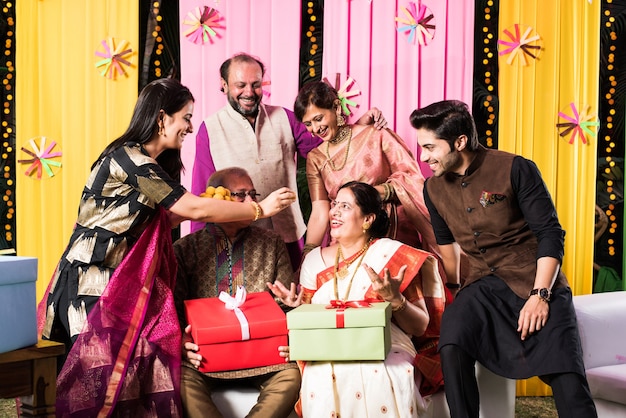 Familia india multigeneracional comiendo dulces mientras celebra un festival u ocasión vestida con ropa tradicional, sentada en un sofá o sofá