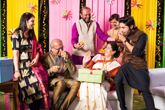 Familia india multigeneracional comiendo dulces mientras celebra un festival u ocasión vestida con ropa tradicional, sentada en un sofá o sofá