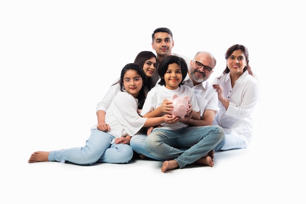 Familia india multigeneracional de seis personas sosteniendo una alcancía mientras usa ropa blanca y está de pie contra la pared blanca