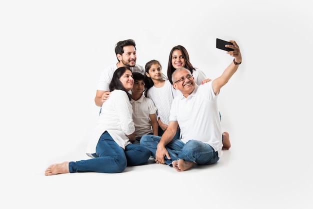 La familia india que toma la foto del selfie con el teléfono inteligente mientras está sentado sobre un fondo blanco incluye 3 generaciones. enfoque selectivo