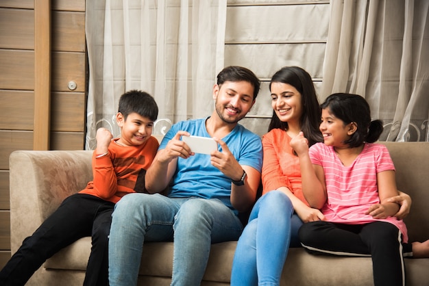 Familia india sentada en el sofá y usando una computadora portátil o tableta con teléfono inteligente viendo películas o navegando por internet