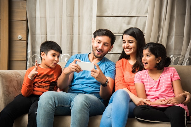 Família indiana sentada no sofá e usando smartphone, laptop ou tablet, assistindo filme ou navegando na internet