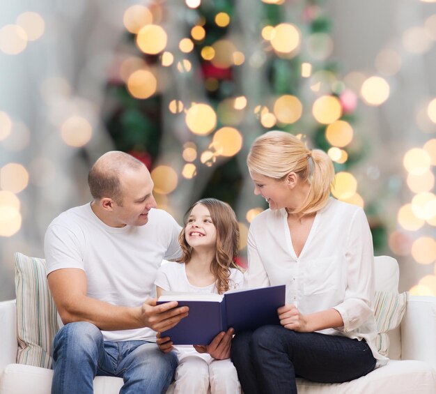 família, infância, feriados e pessoas - sorrindo, mãe, pai e filha lendo livro sobre a sala de estar e o fundo da árvore de natal