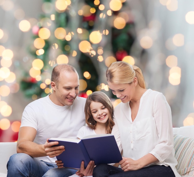 família, infância, feriados e pessoas - sorrindo, mãe, pai e filha lendo livro sobre a sala de estar e o fundo da árvore de natal