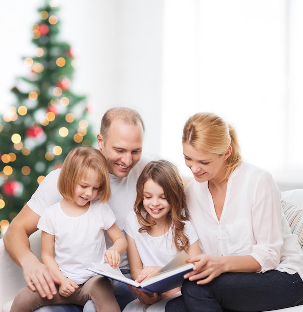 familia, infancia, vacaciones y gente - madre sonriente, padre y niñas pequeñas leyendo un libro sobre la sala de estar y el fondo del árbol de Navidad