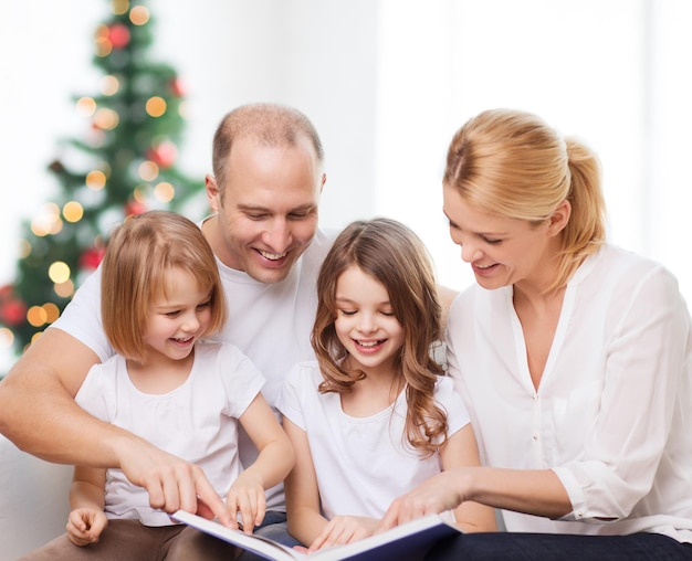 familia, infancia, vacaciones y gente - madre sonriente, padre y niñas pequeñas leyendo un libro sobre la sala de estar y el fondo del árbol de Navidad