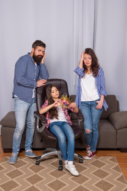 Foto família infeliz de pai e mãe com filha segurando a coroa, sentada na cadeira em casa, fracasso