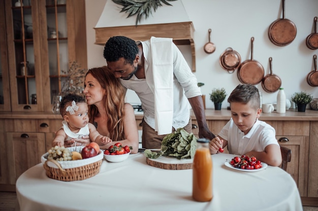 Família inter-racial amigável e sincera com dois filhos passa tempo juntos no café da manhã na cozinha aconchegante