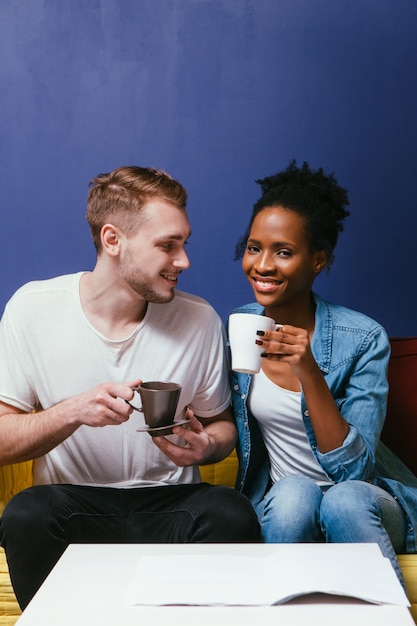 Família interracial jovem e feliz em casa na cena da parede azul, bebendo café ou chá pela manhã