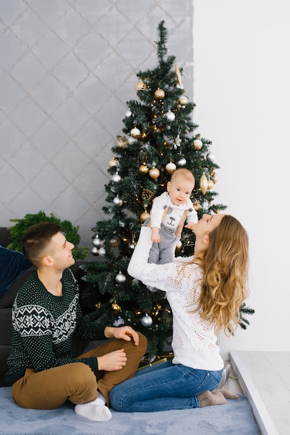 Família jovem amigável: pai, mãe e bebê sentado perto da árvore de natal. mamãe levantou o bebê nos braços e sorriu para ele, o pai olha para eles