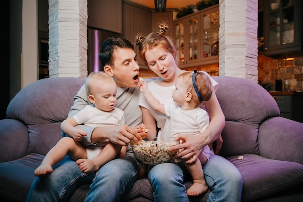 Família jovem com crianças comendo pipoca no sofá em casa
