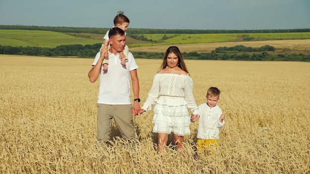 Família jovem com o filho nos ombros do pai e o filho mais velho dando as mãos em um campo de trigo maduro no verão em um dia ensolarado e quente, contra o céu azul no horizonte