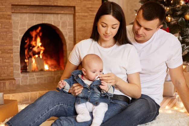 Família jovem comemorando férias de inverno juntos, casal em roupas casuais, sentado no chão com a filha infantil nas mãos, posando no interior com lareira e árvore de natal no fundo.