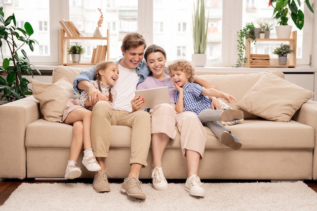 Foto família jovem e alegre e casual com duas crianças e um casal sentado no sofá assistindo a um vídeo engraçado