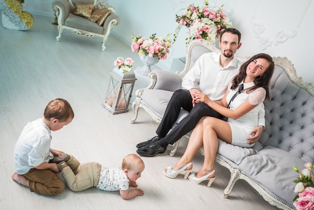 Família jovem e encantadora, um marido e uma esposa amorosos estão sentados em um sofá ao lado de dois filhos bonitos brincando no chão em uma bela sala