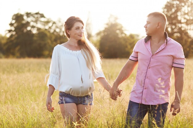 Família jovem feliz, dar as mãos e atravessar o campo de verão, nos olhar com amor. a mulher grávida passa o tempo livre com o marido, respira ar fresco. romance, amor, conceito de relacionamento