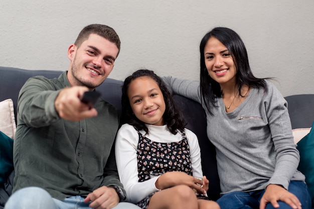 Família jovem inter-racial olhando para a câmera do sofá com o controle remoto. conceito de família assistindo tv em casa.