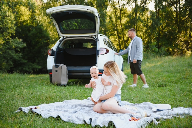 Família jovem três pessoas em roupas brancas fazem piquenique. Filha e pais lindos viajam de carro durante as férias de verão.