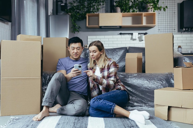 Una familia joven en un apartamento nuevo entre las cajas de cartón sentadas en el suelo y eligiendo felizmente una nueva tienda de muebles en línea usa la aplicación telefónica para comprar