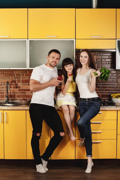 Familia joven atractiva, mamá, papá e hija pequeña posando en una cocina de moda con verduras