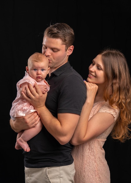 Una familia joven con un bebé en sus brazos Papá besa a la madre recién nacida mira con ternura La niña está vestida con ropa ligera el chico está en una camiseta negra sobre un fondo negro Disparos en el estudio