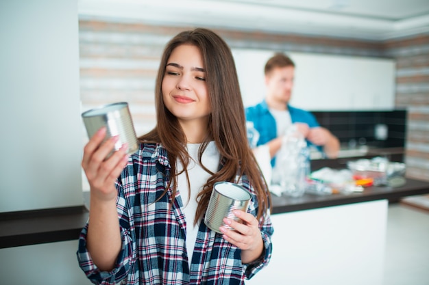 Una familia joven clasifica los materiales en la cocina para reciclarlos. Mujer joven que sostiene las latas viejas en sus manos para la comida. Joven en la pared ordena la basura