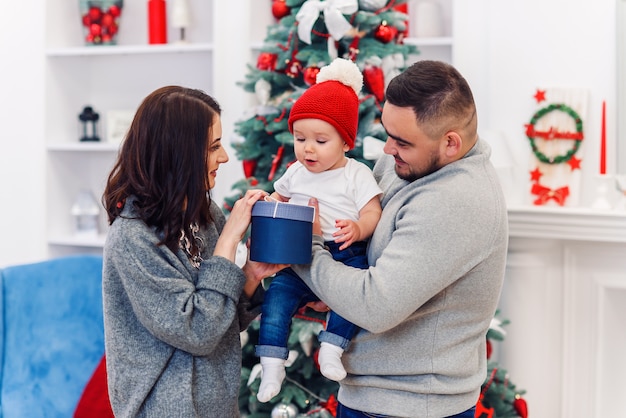 Familia joven le da la primera caja de regalo de Navidad a su hijo recién nacido. El niño pequeño recibe su primer regalo de Navidad.