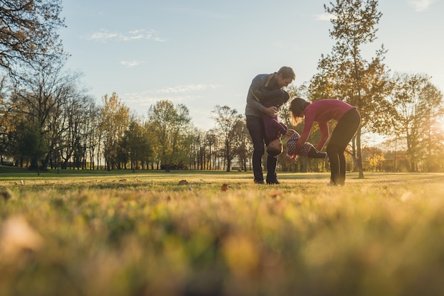 Foto familia joven con dos niños divirtiéndose en un parque en un día soleado de otoño mientras los padres levantan y juegan con sus hijos.