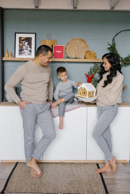 Una familia joven y elegante con un niño pequeño está de pie en su cocina, decorada para Navidad, y admira una casa de pan de jengibre preparada con sus propias manos.