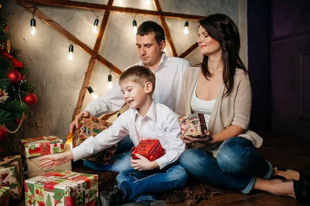 Familia joven feliz en adornos navideños, mamá, papá y niño cerca del árbol de Navidad con regalos cerca