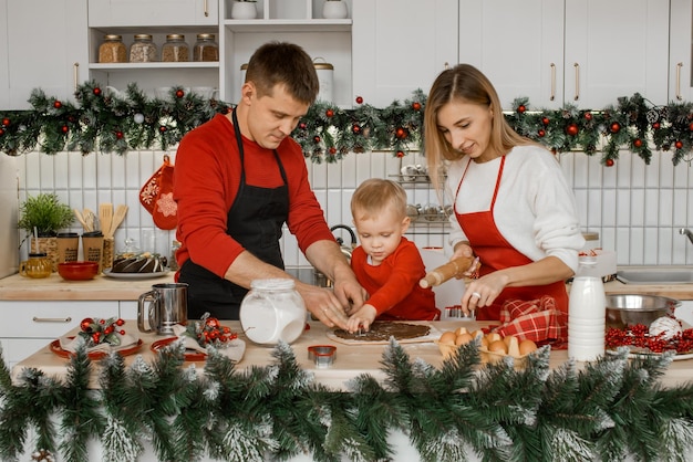 La familia joven feliz en la cocina festiva está muy ocupada cocinando la masa en una mesa blanca Madre y padre viendo a su adorable hijito cortar de la masa Galletas de Navidad