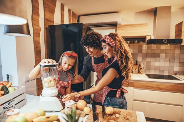 La familia joven feliz está preparando comida saludable en la cocina. Madre, padre e hija están poniendo rebanadas de fruta en la licuadora y haciendo un batido de frutas.