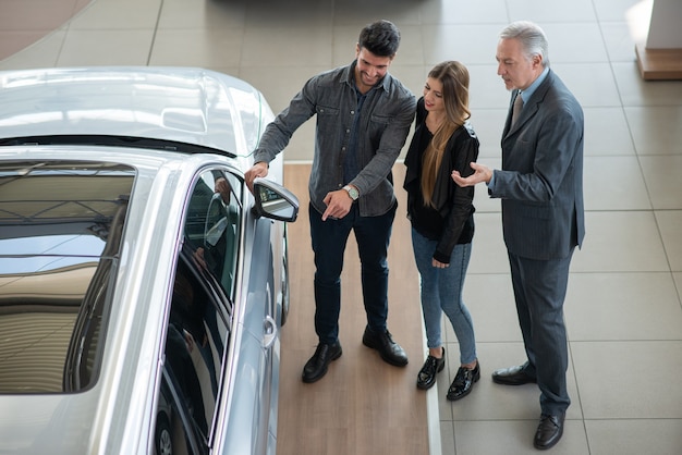Familia joven feliz hablando con el vendedor y eligiendo su nuevo coche en una sala de exposición. Vista desde arriba