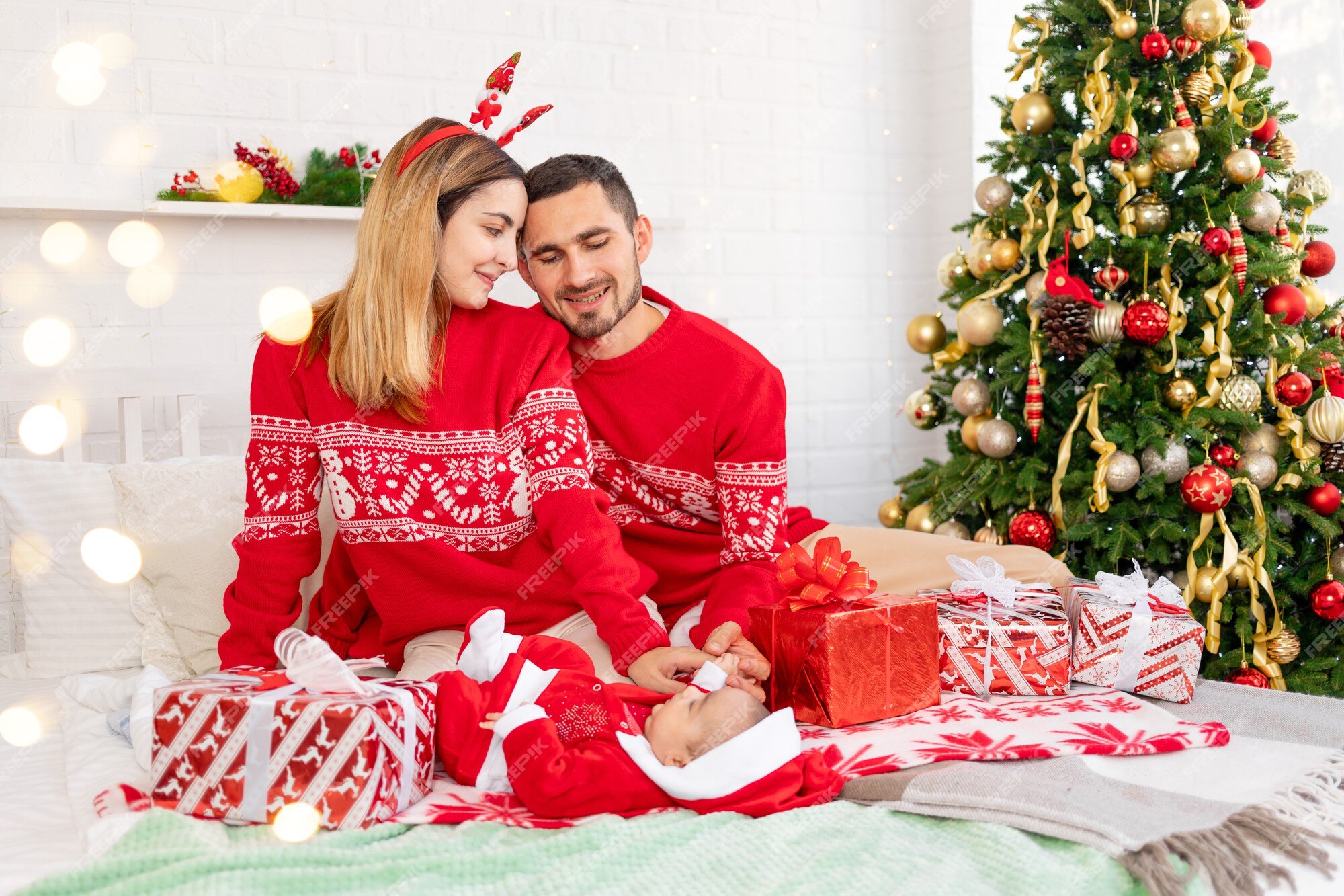 Una familia joven feliz, mamá, papá y bebé en suéteres rojos debajo del árbol de navidad en la cama en casa, celebran el año nuevo o la navidad regocijándose en el regalo Una familia joven feliz, mamá, papá y bebé en suéteres rojos debajo del árbol de navidad en la cama en casa, celebran el año nuevo o la navidad regocijándose en el regalo