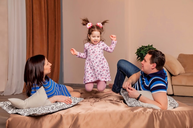 Familia joven feliz con una niña de tres años sentada en la cama y jugando lindo. Encantadora madre embarazada, padre e hija en casa y jugar. Momentos familiares del bebé atmosférico. Copia espacio