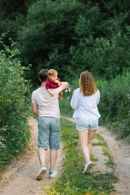 Una familia joven feliz con su hijo en brazos camina por un sendero forestal y disfruta del clima de verano