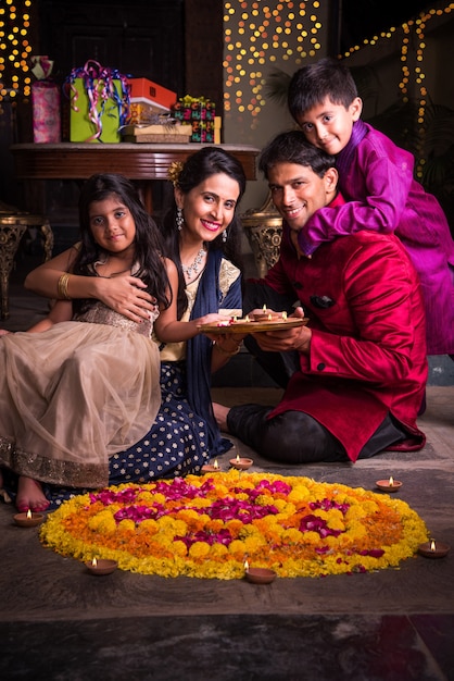 Familia joven india haciendo flor Rangoli o arreglando diyas para la noche del festival de diwali en el porche con regalos y bokeh de fondo