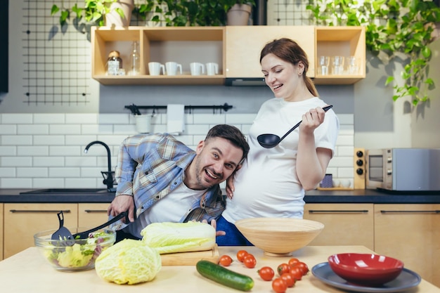 Una familia joven, una mujer embarazada y un hombre, preparan comida saludable y deliciosa juntos en la cocina. Diviértanse juntos. El esposo ayudará a su esposa a preparar la cena.