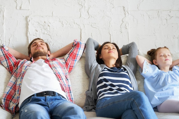 Familia joven relajada descansando y soñando con un nuevo hogar en un cómodo sofá juntos en casa, los padres jóvenes felices con la pequeña hija se relajan disfrutando de la siesta, relajándose o meditando.