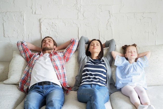 Familia joven relajada descansando y soñando con un nuevo hogar en un cómodo sofá juntos en casa, los padres jóvenes felices con la pequeña hija se relajan disfrutando de la siesta, relajándose o meditando.