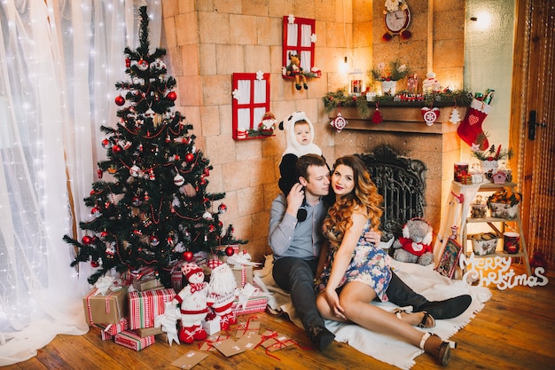Familia joven sentada junto a la chimenea y el árbol de Navidad, hijo sentado en el cuello